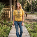 Woman wearing a yellow t-shirt and blue jeans standing on a wooden walkway with hay bales and a horse in the background.